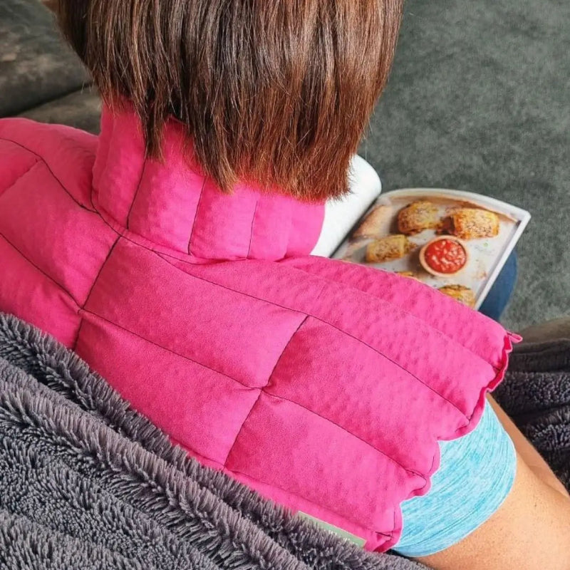Person wearing a pink neck shoulder wheat bag sitting on a couch reading a magazine.